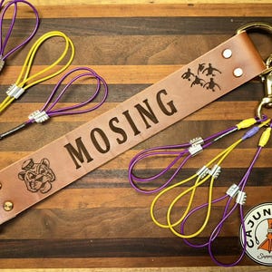 May include: A tan leather duck strap featuring the name MOSING, a tiger mascot logo, and a duck silhouette graphic. It includes gold-tone metal hardware, two large rings, and multiple purple and yellow cord loops with silver-tone metal crimps.