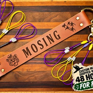 May include: A tan leather strap featuring the text MOSING, a tiger mascot graphic, and a duck silhouette. It includes gold-toned metal hardware and multiple purple and yellow cord loops. The text CUSTOM MADE & SHIPPED WITHIN 48 HOURS FOR FREE is visible.