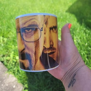 May include: White ceramic mug featuring a photograph of two men's faces. The men are wearing glasses and have facial hair. The mug is being held by a hand with a tattoo on the wrist.