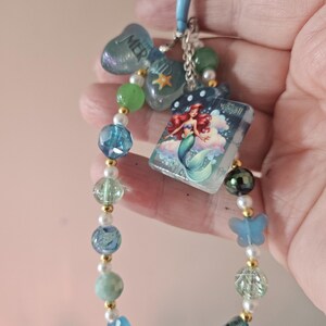 Pode incluir: Um chaveiro de contas com tema de sereia. Apresenta um fecho azul, um la&ccedil;o com a palavra "MERMAID" e um berloque com uma ilustra&ccedil;&atilde;o de sereia. As contas s&atilde;o em tons de azul, verde e branco, com detalhes dourados.