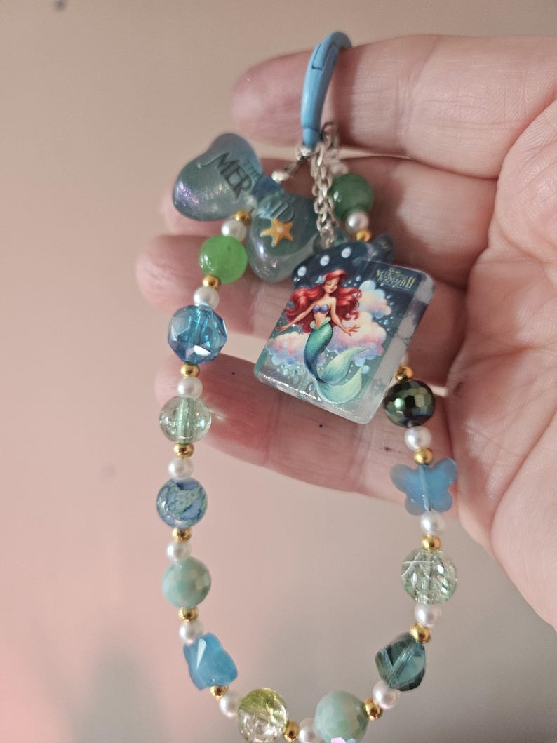 Pode incluir: Um chaveiro de contas com tema de sereia. Apresenta um fecho azul, um la&ccedil;o com a palavra "MERMAID" e um berloque com uma ilustra&ccedil;&atilde;o de sereia. As contas s&atilde;o em tons de azul, verde e branco, com detalhes dourados.