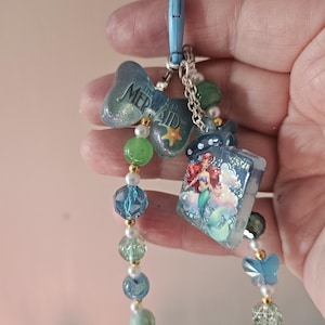 Pode incluir: Uma pulseira de contas azuis e verdes com fecho de lagosta. A pulseira apresenta berloques com o texto "The Mermaid", uma estrela e uma ilustra&ccedil;&atilde;o de sereia. As contas s&atilde;o de v&aacute;rias formas e tamanhos, com detalhes dourados.