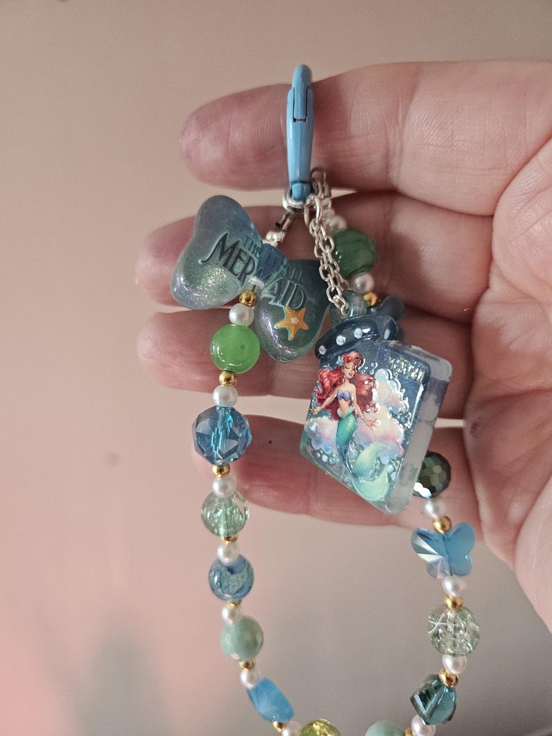 Pode incluir: Uma pulseira de contas azuis e verdes com fecho de lagosta. A pulseira apresenta berloques com o texto "The Mermaid", uma estrela e uma ilustra&ccedil;&atilde;o de sereia. As contas s&atilde;o de v&aacute;rias formas e tamanhos, com detalhes dourados.