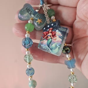 Pode incluir: Um chaveiro azul com tema de sereia. O chaveiro apresenta um pingente com o texto "MERMAID", um pequeno livro com uma ilustra&ccedil;&atilde;o de Ariel e uma corrente de contas azuis, verdes e peroladas. Um pingente de borboleta azul tamb&eacute;m est&aacute; inclu&iacute;do.
