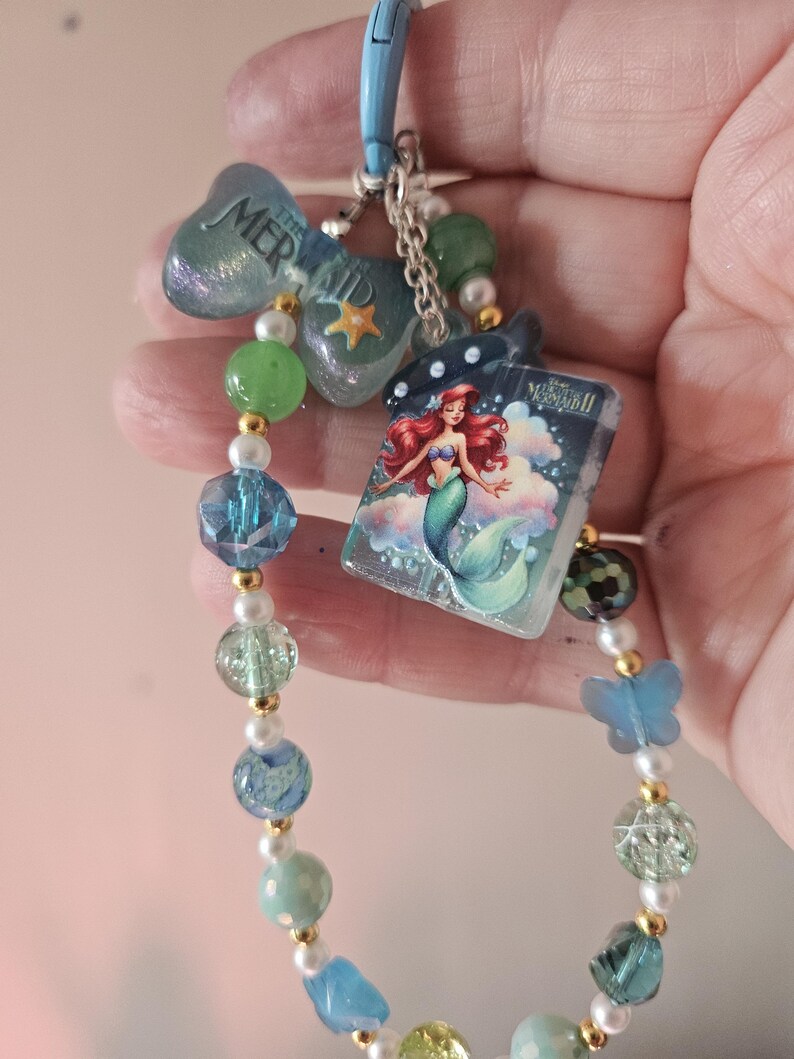 Pode incluir: Um chaveiro azul com tema de sereia. O chaveiro apresenta um pingente com o texto "MERMAID", um pequeno livro com uma ilustra&ccedil;&atilde;o de Ariel e uma corrente de contas azuis, verdes e peroladas. Um pingente de borboleta azul tamb&eacute;m est&aacute; inclu&iacute;do.