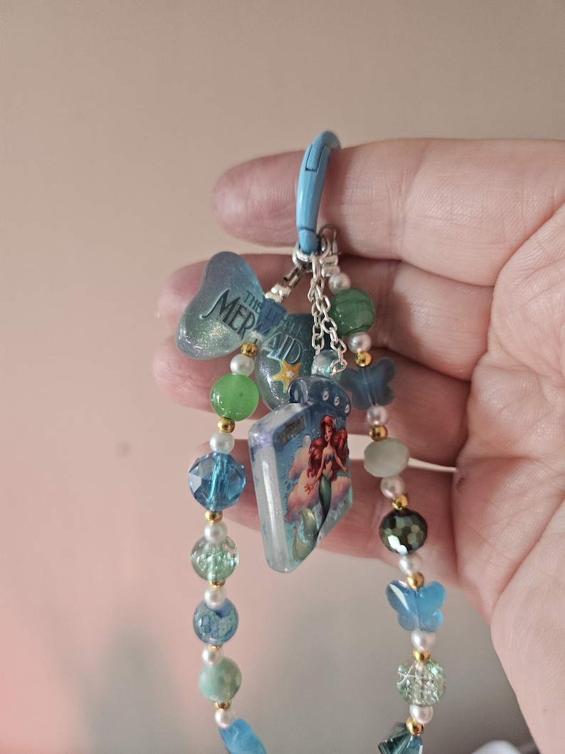Pode incluir: Um chaveiro azul com uma variedade de pingentes, incluindo um livro com uma ilustra&ccedil;&atilde;o de sereia, uma borboleta e detalhes de contas. O chaveiro tem um fecho azul e &eacute; adornado com p&eacute;rolas e contas douradas.