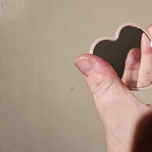 May include: A small, heart-shaped mirror with a pale pink frame and a black reflective surface. The mirror is held up against a neutral background.