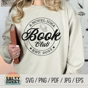 K&ouml;nnte beinhalten: Ein wei&szlig;es Sweatshirt mit einem schwarzen Kreisdesign, auf dem "A Novel Idea Book Club Est. 2023" steht und sich eine Stapel B&uuml;cher im Kreis befindet.