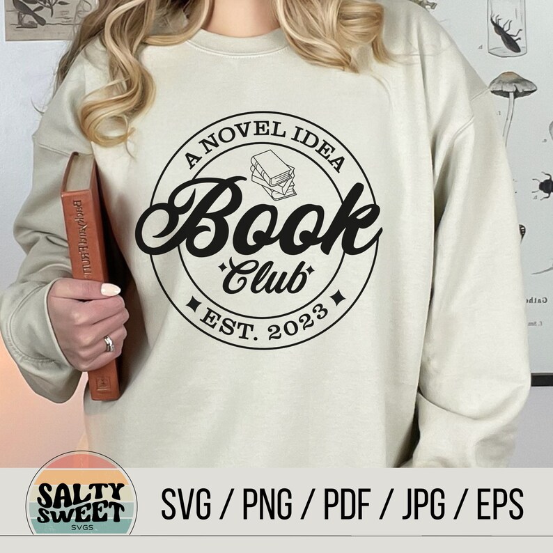 K&ouml;nnte beinhalten: Ein wei&szlig;es Sweatshirt mit einem schwarzen Kreisdesign, auf dem "A Novel Idea Book Club Est. 2023" steht und sich eine Stapel B&uuml;cher im Kreis befindet.