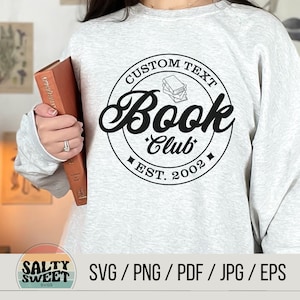 K&ouml;nnte beinhalten: Ein graues Sweatshirt mit einer schwarz-wei&szlig;en Grafik eines Buchclub-Logos. Das Logo zeigt einen Stapel B&uuml;cher, die Worte "Custom Text Book Club" und "Est. 2002".