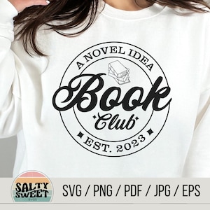 K&ouml;nnte beinhalten: Ein wei&szlig;es Sweatshirt mit einem schwarzen Kreisdesign, auf dem "A Novel Idea Book Club Est. 2023" steht und sich eine Stapel B&uuml;cher im Kreis befindet.