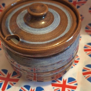 May include: A brown and blue ceramic sugar bowl with a lid. The lid has a concentric circle design with blue stripes. The bowl has a blue and white design.