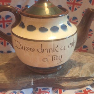 May include: A cream-coloured ceramic teapot with a brown handle and spout, and a green and brown lid. The teapot is decorated with blue dots and the words "Duee drink a cup a Tay". It sits on a piece of wood, against a Union Jack heart backdrop.