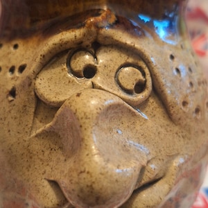 May include: A quirky, handmade ceramic mug featuring a sculpted face. The mug is mainly tan with brown detailing and a glossy finish. The face has large eyes and a prominent nose. The mug is approximately 13 cm tall.