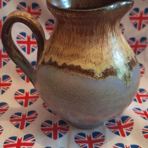 May include: A ceramic jug with a handle, featuring a rounded body and a flared rim. The jug is glazed in shades of brown, yellow, and blue-grey. The background is a fabric printed with heart-shaped Union Jacks.
