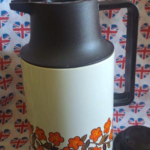 Könnte beinhalten: Eine weiße und braune Isolierkanne im Vintage-Stil mit schwarzem Griff und Deckel. Die Kanne hat ein orangefarbenes Blumendesign am Boden. Der Hintergrund ist ein Stoff mit herzförmigen Union-Jack-Flaggen.