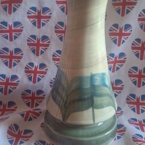 May include: A cream-coloured ceramic vase with a flared top and a wide base. The vase has a painted design of blue and green flags. The background is a white fabric with a repeating pattern of hearts in the colours of the Union Jack.