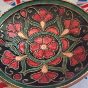 May include: A round, decorative bowl with a floral design. The bowl has a black background with red and pink flowers, green accents, and copper-coloured handles. The bowl is approximately 15 cm in diameter.