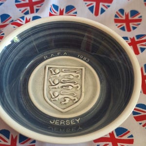 May include: A ceramic bowl with a blue interior and white rim, featuring a crest and the word "Jersey". The bowl is set against a white fabric patterned with heart-shaped Union Jack flags.