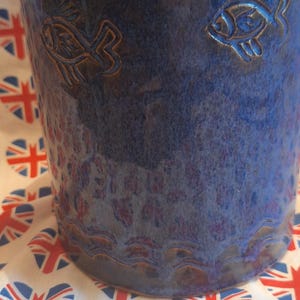 May include: A blue ceramic plant pot with a textured finish and embossed fish motifs. The pot has a wavy rim and sits on fabric printed with a heart-shaped Union Jack design.
