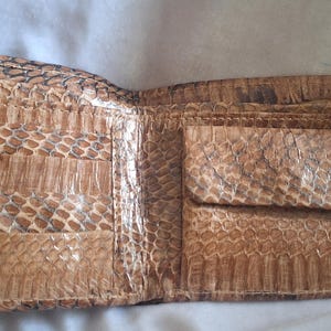 May include: A bi-fold wallet crafted from light brown snakeskin with a distinct scale pattern. The interior features a horizontal bill compartment and a small flap-covered coin pocket on the right side, all finished in the same textured snakeskin material.