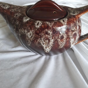 May include: A brown ceramic teapot with a lid. The teapot features a speckled pattern with white accents. The lid is a darker shade of brown. The teapot has a handle and a spout. The teapot is on a white surface.