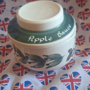 May include: A white ceramic apple sauce pot with a lid, featuring green trim and the words "Apple Sauce". The pot is decorated with green leaves and sits on a fabric with heart-shaped Union Jack patterns.