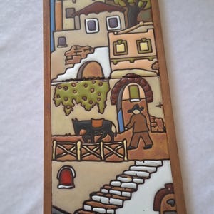 May include: A vertical rectangular wooden frame containing two stacked ceramic tiles featuring a colourful hand-painted scene of a Mediterranean village with houses, a person leading a donkey, stone stairs, and a figure carrying a jug, signed AT.