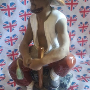 May include: A ceramic figurine depicting a seated Asian man in traditional attire, including a conical hat. The figure is painted in brown, white, and red. He holds a wooden tool. The figurine sits on a rock-like base.