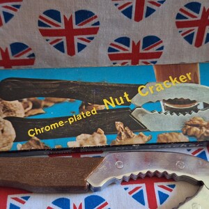 May include: A chrome-plated nutcracker with a wooden handle and a box with the words "Nut Cracker" on it. The box is blue with an orange stripe. The background is a white cloth with red, white, and blue heart-shaped Union Jack patterns.