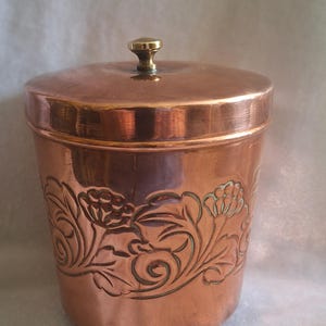 May include: A cylindrical copper canister with a polished finish, featuring an embossed floral pattern around the centre. The container has a matching lid with a small, central brass-coloured knob and rests on three small, rounded feet.