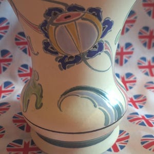 May include: A cream-coloured ceramic vase featuring a floral design in blue, yellow, and pink. The vase has a wide top and a rounded base. The background is a fabric with heart-shaped Union Jack patterns.