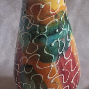 May include: A brightly coloured ceramic vase with a distinctive pattern. The vase displays a vibrant blend of colours: yellow, orange, red, green, and blue, with white squiggly lines. It has a rounded body and a slightly flared opening.