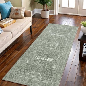 May include: A rectangular, sage green runner rug with an ornate, floral pattern. The rug features a central medallion design and a detailed border. It is placed on a dark wood floor, near a beige sofa and a small side table.