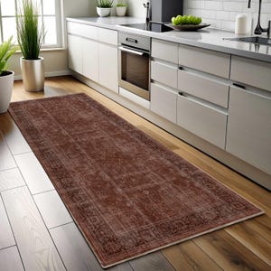 May include: A rectangular, distressed-look rug in shades of brown and red, placed on a wooden floor in a kitchen setting. The rug features a detailed border and a faded, vintage appearance, adding a touch of warmth and style to the space.