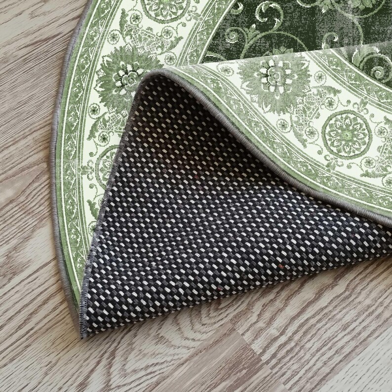 Pode incluir: Uma sec&ccedil;&atilde;o de um tapete redondo com um design floral em tons de verde e creme. A parte inferior do tapete &eacute; preta com um padr&atilde;o de pontos brancos, e a borda &eacute; cinza. O tapete est&aacute; parcialmente enrolado, revelando o verso.