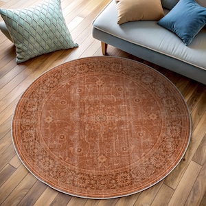 May include: A round, rust-colored rug with an intricate floral pattern. The rug has a distressed, vintage look with a detailed border. The rug is placed on a hardwood floor, with a sofa and pillows in the background.