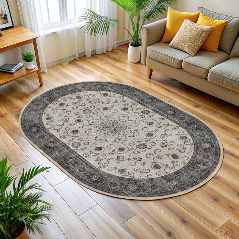 Pode incluir: Um tapete oval com um centro creme com um padr&atilde;o floral em tons de castanho e cinza. O tapete tem uma borda cinza escuro com um design circular repetitivo. Est&aacute; colocado sobre um ch&atilde;o de madeira numa sala de estar.