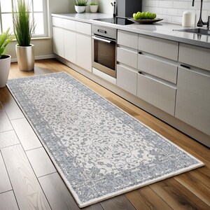 Puede incluir: Una alfombra de pasillo rectangular con un diseño floral en blanco roto y gris. La alfombra tiene un borde gris y está colocada sobre un suelo de madera en una cocina. La alfombra es un elemento decorativo.