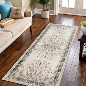 May include: A rectangular beige rug with an ornate floral pattern and a dark border. The rug is placed on a dark wood floor, and is in a living room setting.