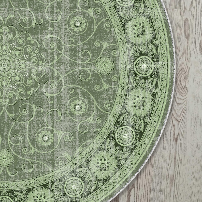May include: A round, olive green rug with a distressed, vintage look. The rug features a central floral medallion design with scrolling vines and flowers. A darker green border with floral medallions frames the central design. The rug is placed on a light wood floor.
