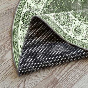 May include: A section of a round rug with a floral design in shades of green and cream. The rug's underside is black with a white dotted pattern. The rug is partially rolled up, revealing the backing.