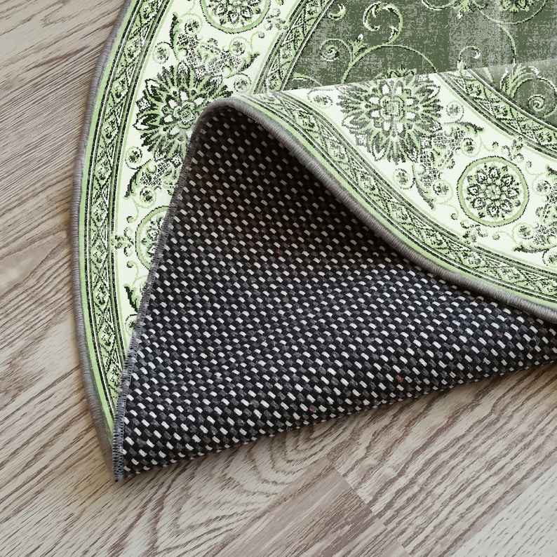 May include: A section of a round rug with a floral design in shades of green and cream. The rug's underside is black with a white dotted pattern. The rug is partially rolled up, revealing the backing.
