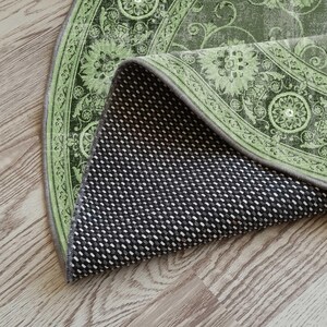 May include: A section of a round rug with a floral design in shades of green and gray. The rug's underside is visible, showing a black and white dotted pattern. The rug is partially rolled up, revealing its construction and texture.