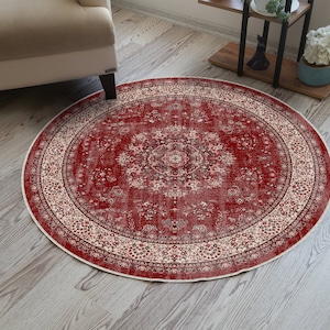 Può includere: Un tappeto rotondo rosso con un intricato motivo floreale. Il tappeto presenta un design a medaglione centrale e un bordo color crema con motivi floreali. È posato su un pavimento in legno chiaro, con una gamba di poltrona beige visibile. Diametro: circa 150 cm.