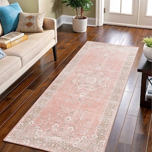 May include: A rectangular rug with a distressed appearance, featuring a faded pink central area and a beige border. The rug displays a central medallion and floral designs. It's positioned on a dark wood floor, suitable for a living room or hallway.