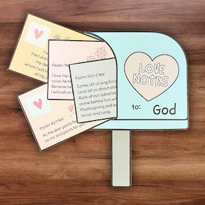 Puede incluir: Un buzón azul con una abertura en forma de corazón y las palabras "Love Notes to: God" en la parte delantera. Hay cuatro tarjetas dentro del buzón con versículos bíblicos impresos en ellas.