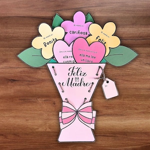 Op de afbeelding: Een handgemaakt papieren boeket voor Moederdag. Het boeket is roze met een strik en heeft bloem- en hartvormige uitsparingen met Spaanse tekst. De tekst bevat zinnen als "Feliz Día de la Madre" en "Mi mamá es bonita."