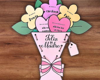May include: A handmade paper bouquet for Mother's Day. The bouquet is pink with a bow and features flower and heart-shaped cutouts with Spanish text. The text includes phrases like "Feliz Día de la Madre" and "Mi mamá es bonita."