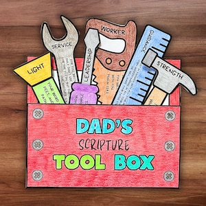 May include: A red toolbox with the words "DAD'S SCRIPTURE TOOL BOX" in blue and green. Inside are paper cutouts of tools labeled "SERVICE," "LEADERSHIP," "WORKER," "GUIDANCE," and "STRENGTH." The tools are on a wooden surface.